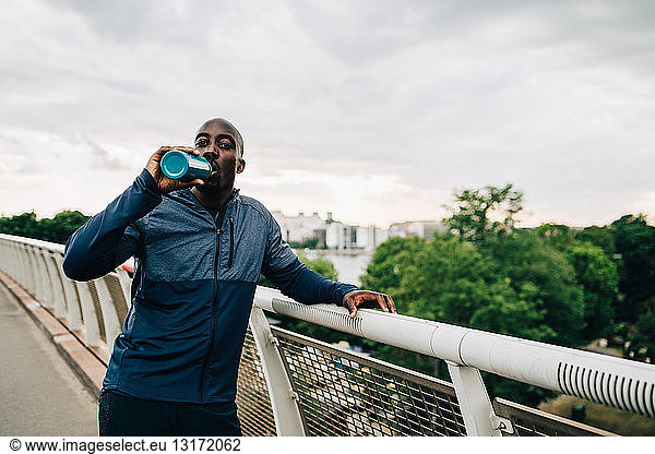 Portrait of thirsty sportsman drinking from bottle while standing on footbridge against sky