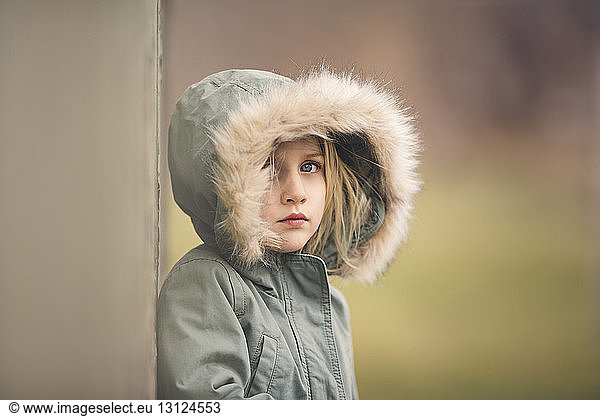 Portrait of thoughtful girl wearing fur hood standing against wall