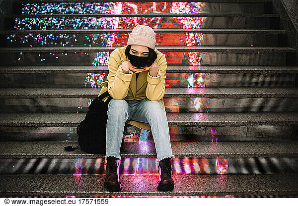 Portrait of thoughtful young woman wearing face mask sitting on steps