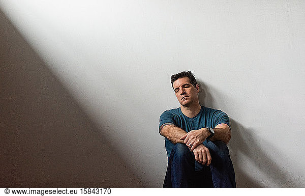 Portrait of tired man leaning against white wall with his eeditorial closed.