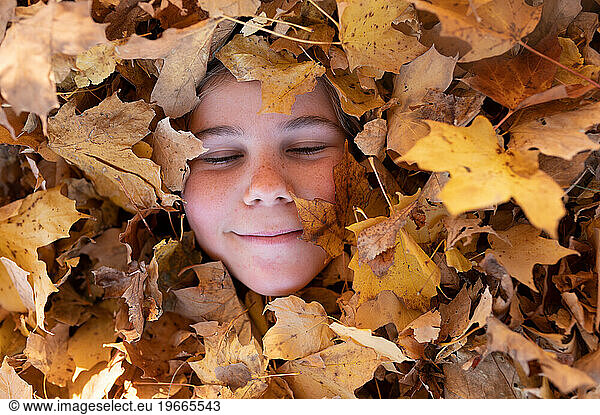 Portrait of tween with eyes closed buried in yellow autumn leaves