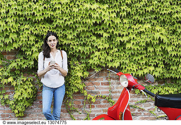 Portrait of woman holding smart phone while standing by motor scooter against ivy wall