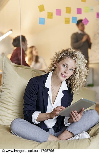 Portrait of woman in office using tablet in bean bag with meeting in background