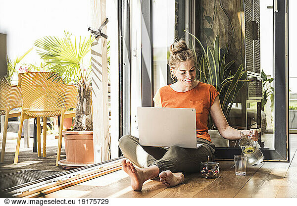 Portrait of woman sitting by open balcony with laptop pouring water