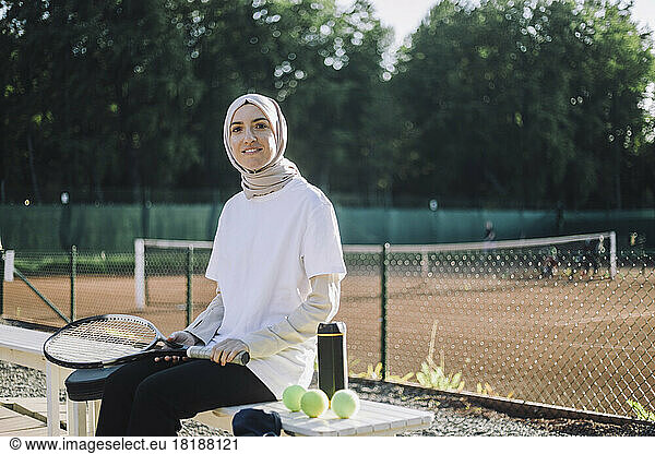 Portrait of woman wearing headscarf holding tennis racket while sitting on bench at sports court