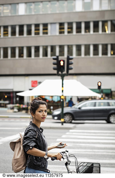 Portrait of woman with bicycle holding mobile phone while standing on city street