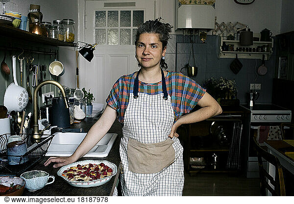 Portrait of woman with hand on hip standing by pie on kitchen counter at home