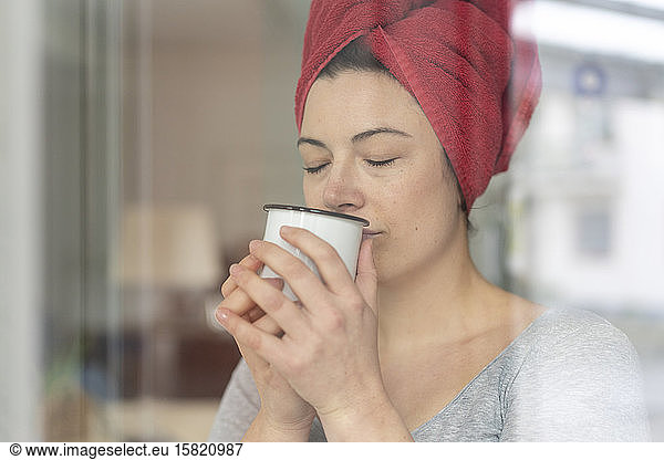Portrait of woman with head wrapped in a towel drinking a coffee behind windowpane