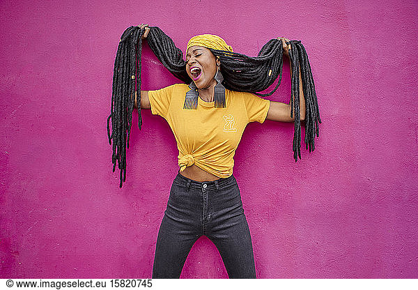 Portrait of woman with long dreadlocks holding her hair in front of a pink wall