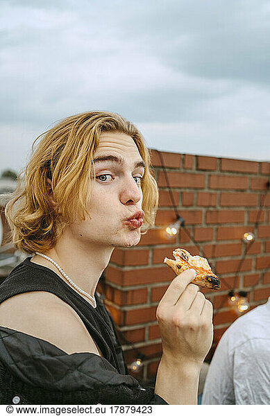 Portrait of young blond man with pizza puckering lips on rooftop