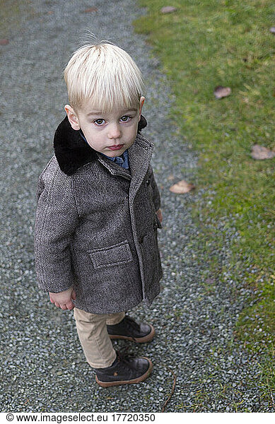 Portrait of young boy with platinum blond hair wearing tweed coat and standing on a gravel trail looking up at the camera; Aldergrove  British Columbia  Canada