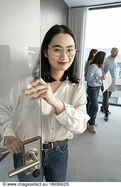 Portrait of young businesswoman opening door with colleagues in background in office