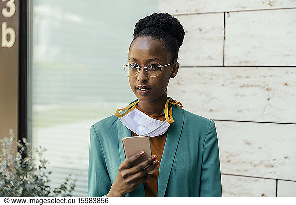 Portrait of young businesswoman with protective mask and smartphone outdoors