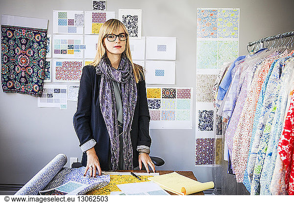 Portrait of young female fashion designer standing by desk in workshop