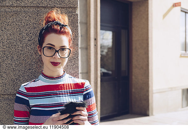 Portrait of young female hipster holding mobile phone while standing against wall