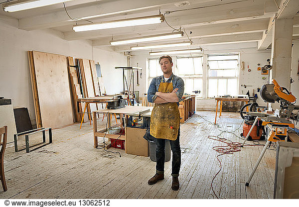 Portrait of young male carpenter standing with arms crossed in workshop