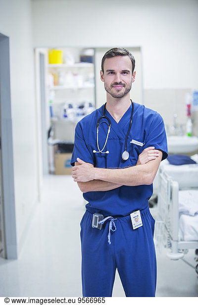 Portrait of young male doctor standing in hospital ward with arms crossed