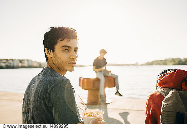 Portrait of young man holding meal in container while sitting on promenade during sunny day