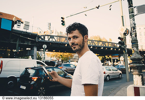 Portrait of young man holding mobile phone while standing on sidewalk in city