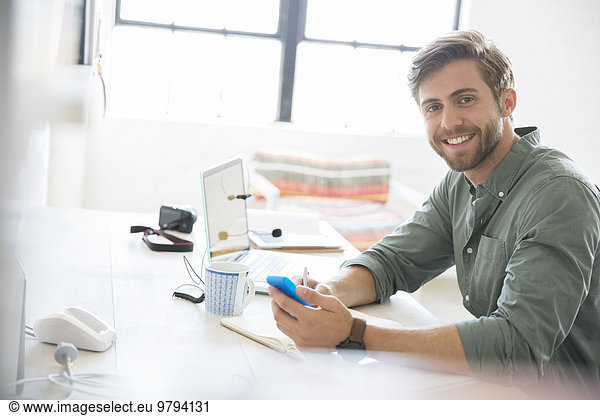 Portrait of young man sitting at desk with mobile phone and laptop