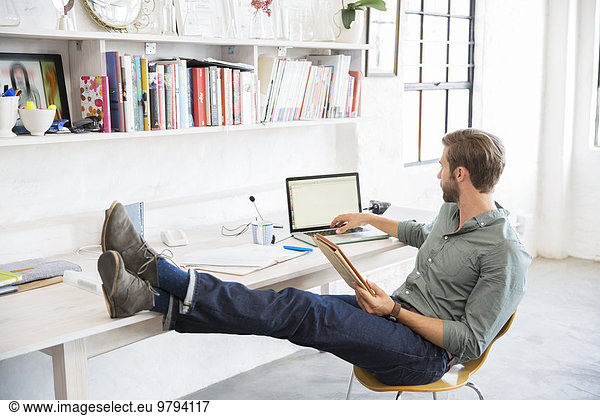 Portrait of young man sitting with legs on desk working with laptop