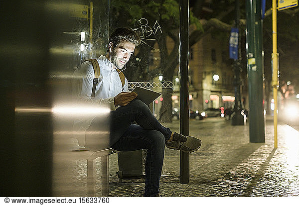 Portrait of young man waiting at bus stop by night using digital tablet and earphones  Lisbon  Portugal