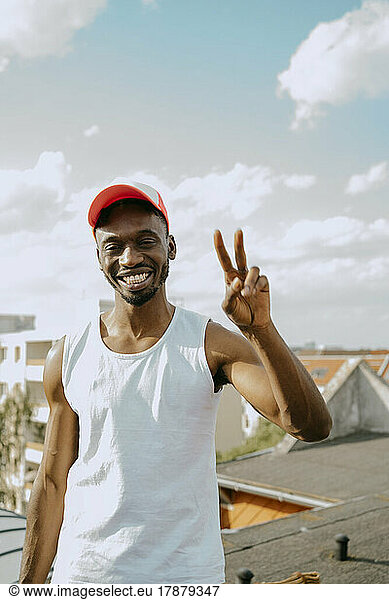 Portrait of young man wearing cap gesturing peace sign against sky on sunny day