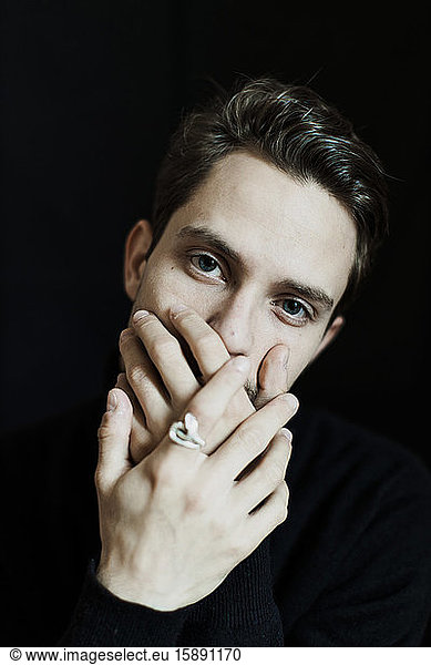 Portrait of young man with hands covering mouth against black background