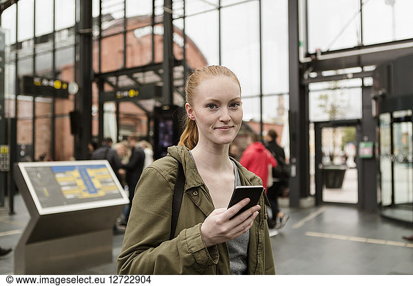 Portrait of young woman holding mobile phone while standing at railroad station