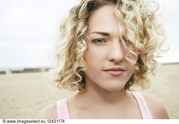 Portrait of young woman with blond curly hair standing on sandy beach  looking at camera.