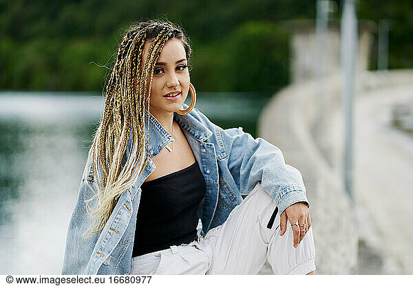 Portrait of young woman with braided hair wearing a denim jacket sitting on a bridge