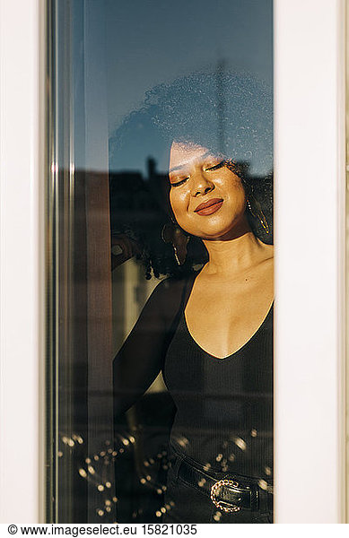 Portrait of young woman with curly hair behind windowpane enjoying the sunshine
