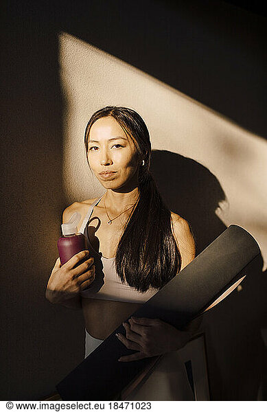 Portrait of young woman with exercise mat and water bottle standing against wall