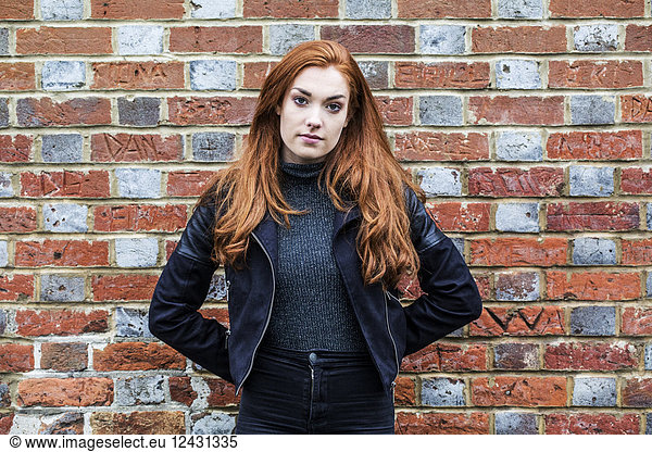 Portrait of young woman with long red hair standing in front of brick wall.