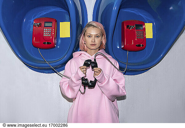 Portrait of young woman with pink hair wearing pink hooded shirt standing in front of telephone booths