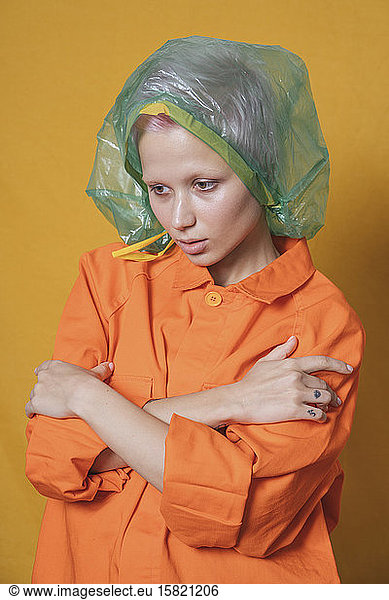 Portrait of young woman with plastic bag on her head wearing orange jacket in front of yellow background