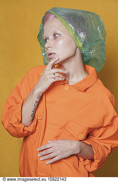 Portrait of young woman with plastic bag on her head wearing orange jacket in front of yellow background