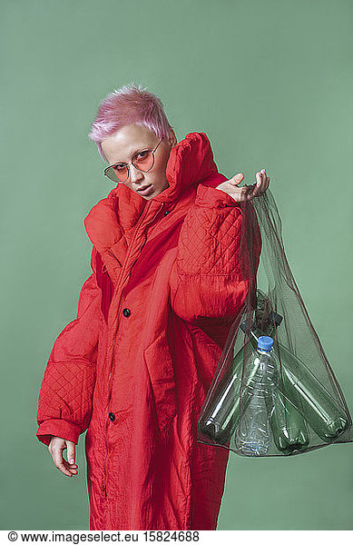 Portrait of young woman with short pink hair wearing red coat holding pouch with plastic bottles