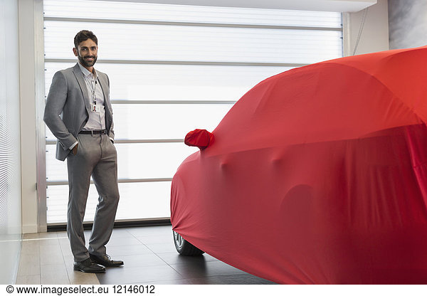 Portrait selbstbewusster Autoverkäufer neben dem gedeckten Auto im Autohaus-Showroom