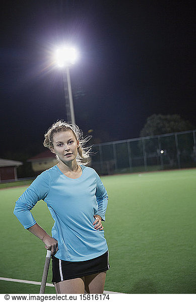 Portrait serious young female field hockey player on field at night