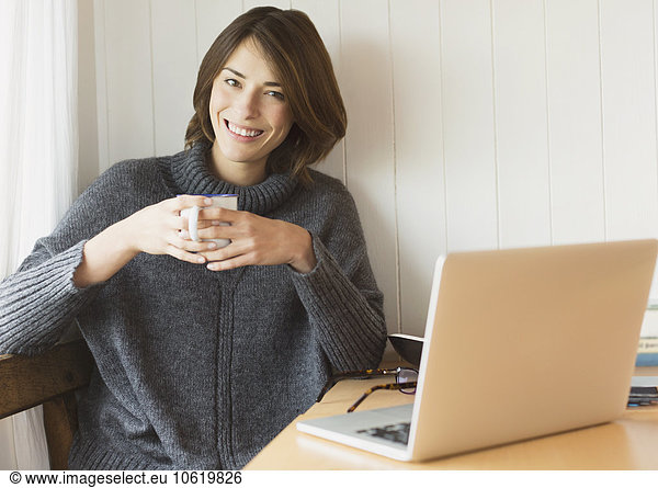 Portrait smiling brunette woman in sweater drinking coffee at laptop