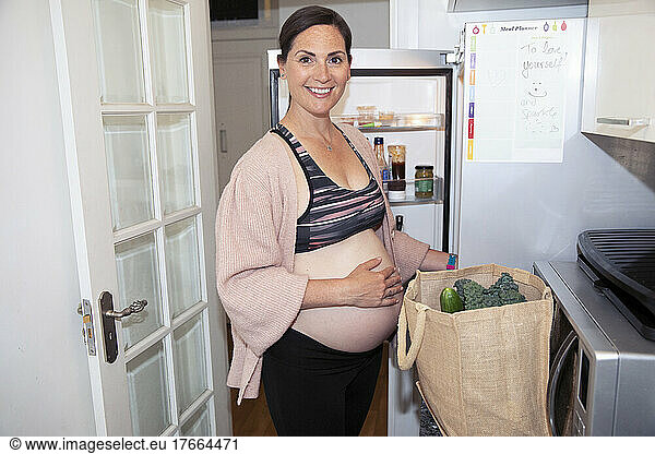 Portrait smiling pregnant woman in sports bra unloading groceries