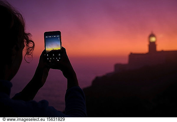 Portugal  Algarve  Über-Schulter-Ansicht einer Person  die in der Morgendämmerung per Smartphone Fotos vom Leuchtturm von Cape Saint Vincent macht