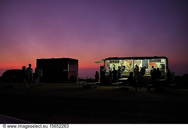 Portugal  Algarve  Purpurner Himmel über Silhouetten von Menschen  die sich in der Morgendämmerung am Konzessionsstand in Cape Saint Vincent entspannen