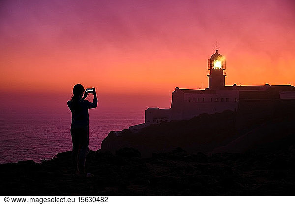 Portugal  Algarve  Silhouette einer Person  die in der Morgendämmerung per Smartphone Fotos vom Leuchtturm von Cape Saint Vincent macht