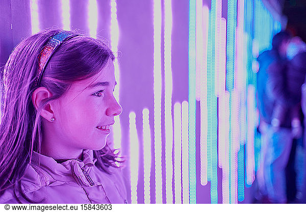 Positive young girl in casual clothes standing near glowing neon wall
