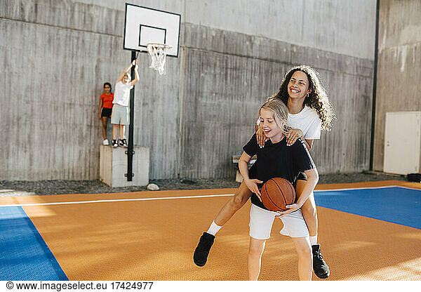 Pre-adolescent girl carrying female friend piggyback at basketball court