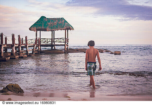 Pre teen boy walking into the ocean by a wooden pier.