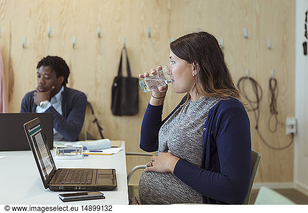 Pregnant businesswoman drinking water while sitting with laptop in meeting