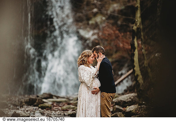 Pregnant couple kissing outside by a waterfall standing on rocks